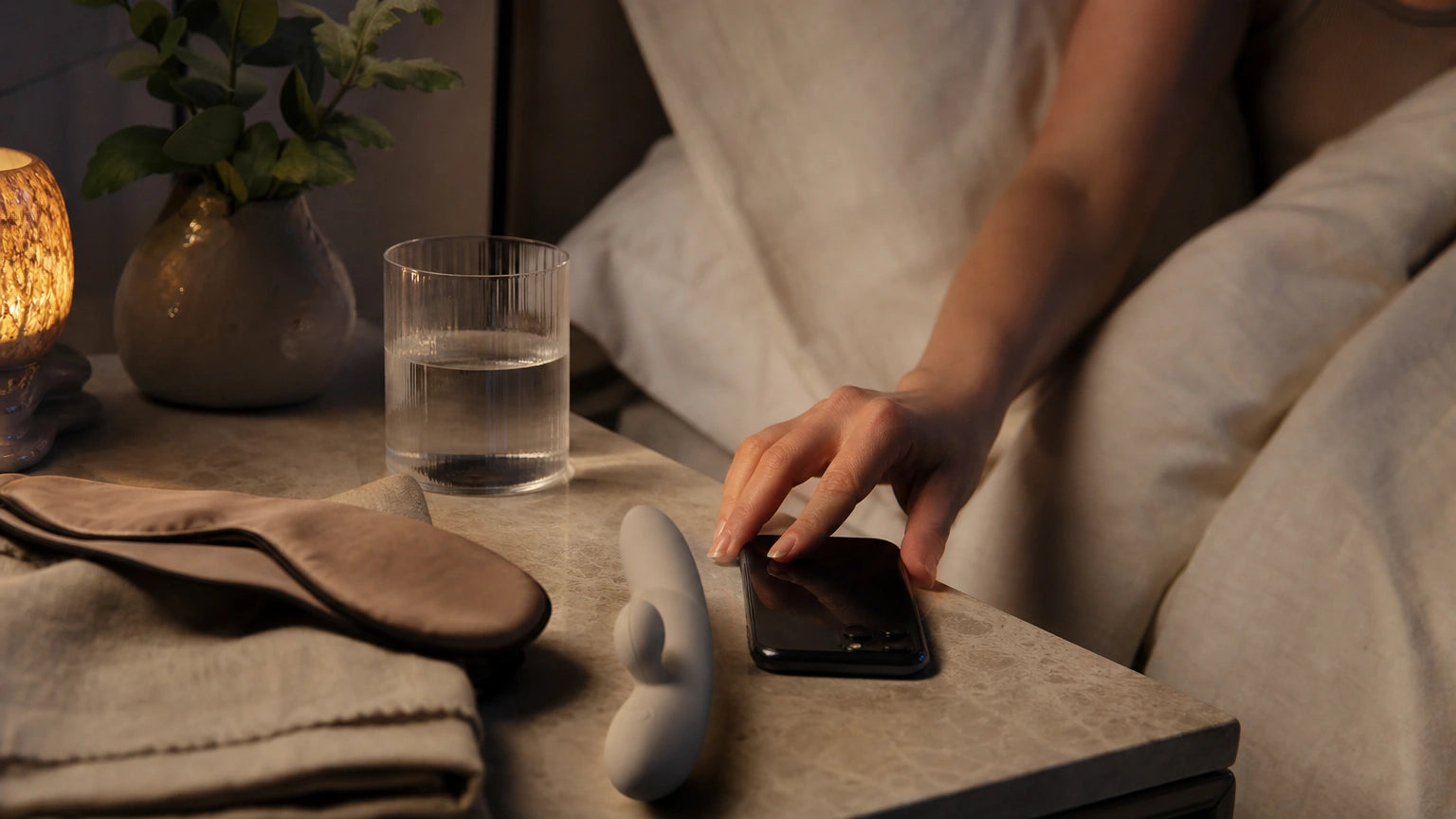 Smartphone placed face down beside a discreet silicone massager on a calm bedside table for an evening stress relief