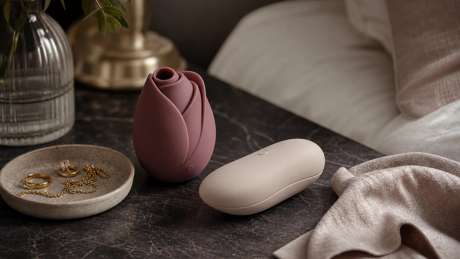 Rose-shaped vibrator and pebble-style silicone massager styled as elegant self-care gifts on a bedroom tabletop