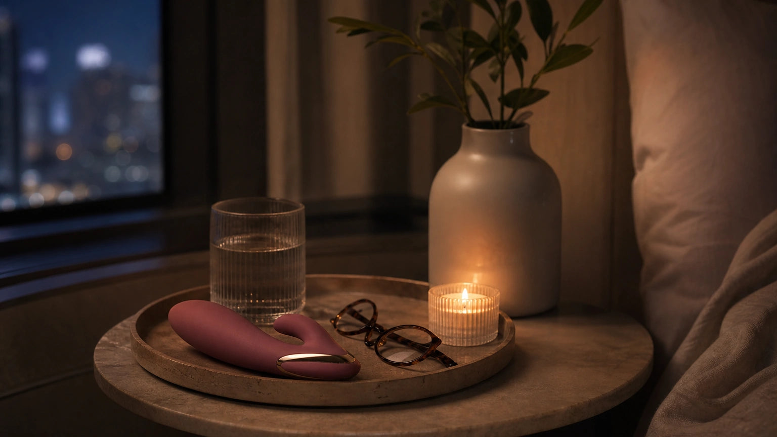 Discreet vibrator on a nightstand tray beside reading glasses, water, and candle in a minimalist bedroom