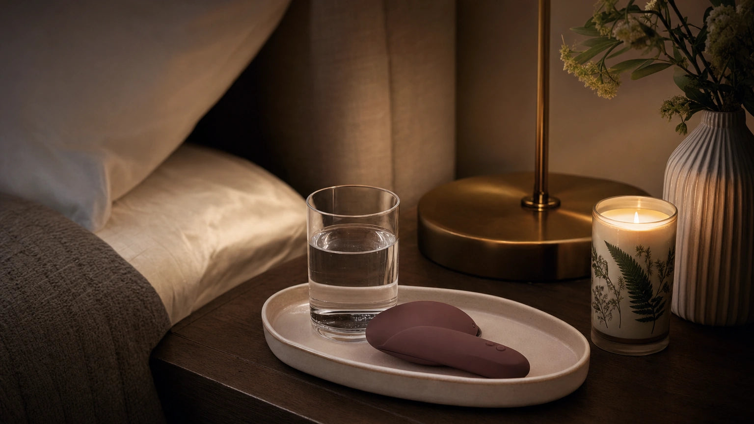 Pebble-shaped vibrator on a ceramic tray with water, lamp, and candle on a calm nightstand